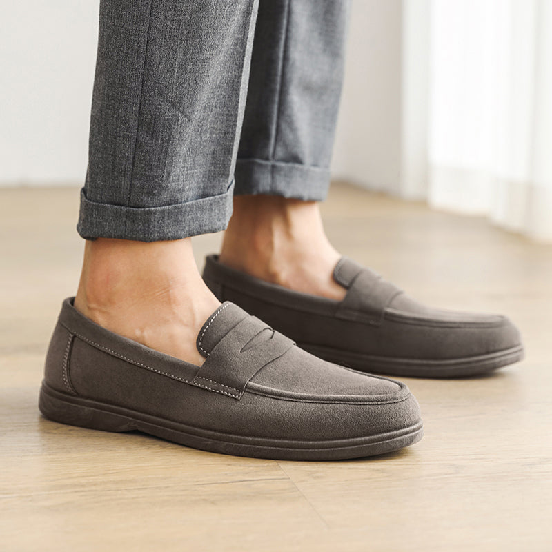 Close-up of a person's feet wearing gray suede loafers with cuffed gray trousers, standing on a wood floor.