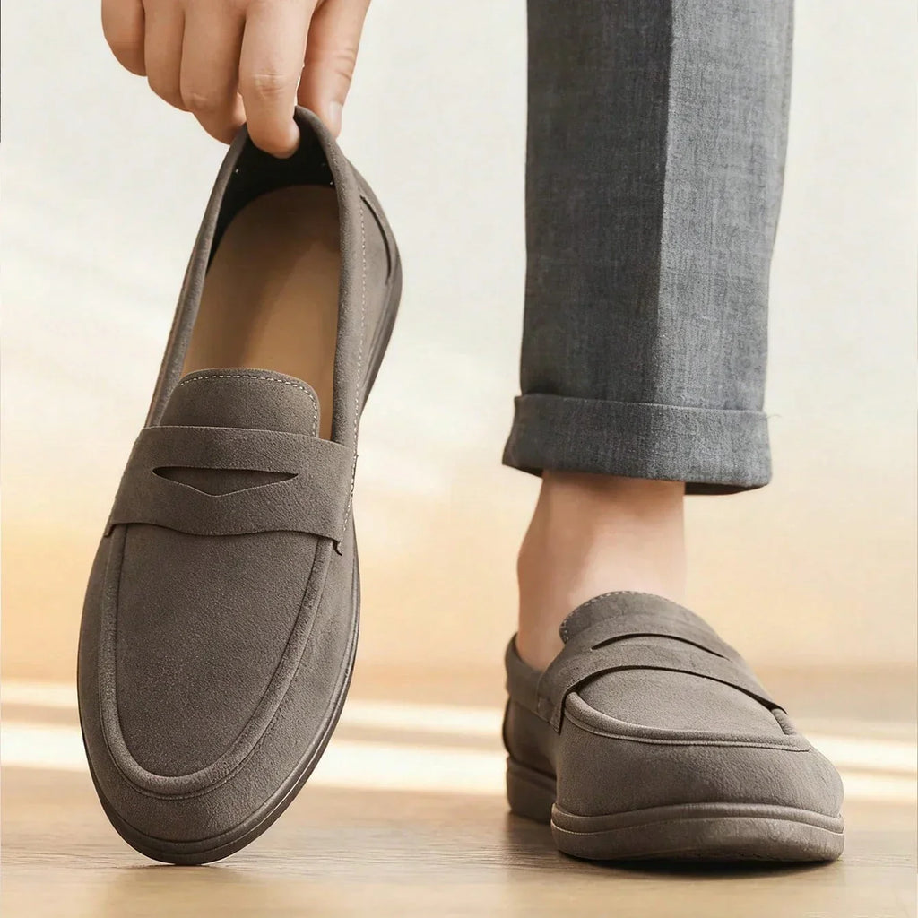 A person holds up one gray suede loafer while wearing the other, cuffed gray trousers visible, warm side lighting.