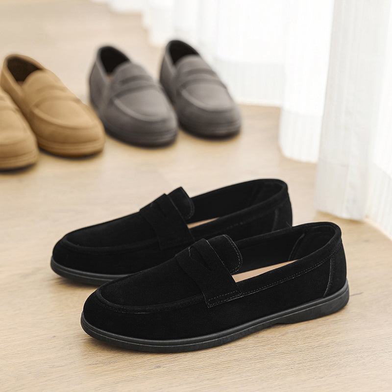 Black suede penny loafers in the foreground with tan and gray suede loafers lined up behind on a wooden floor beside a curtain.