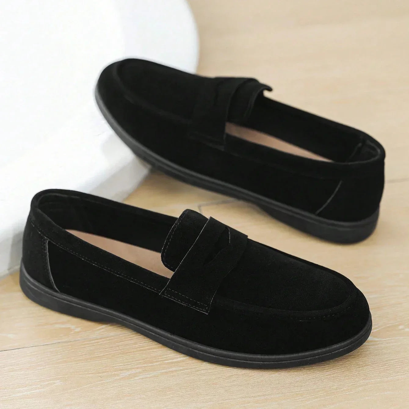 Pair of black suede penny loafers resting on a wood floor against a curved white wall.