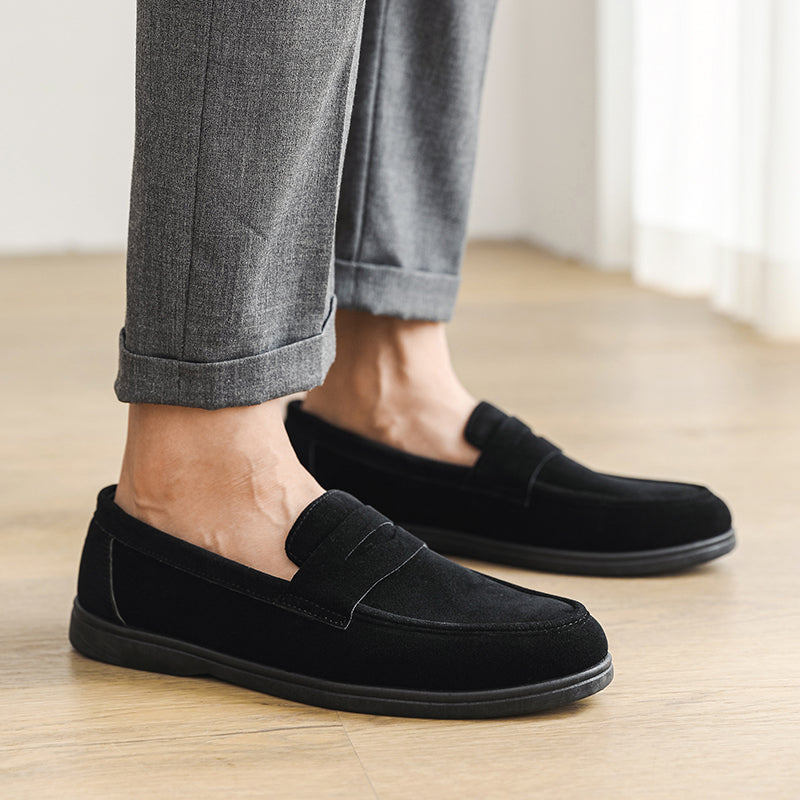 Close-up of a person's feet wearing black suede loafers with cuffed gray trousers, standing on a wood floor.