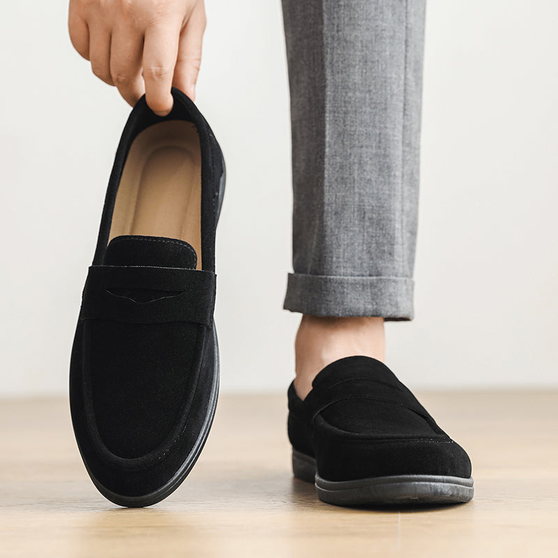 A person holds up one black suede loafer while wearing the other, cuffed gray trousers visible against a plain background.