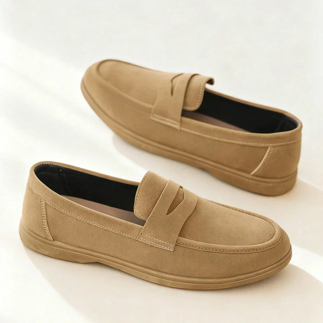 Pair of tan suede penny loafers with stitched apron toe, photographed on a plain white background.