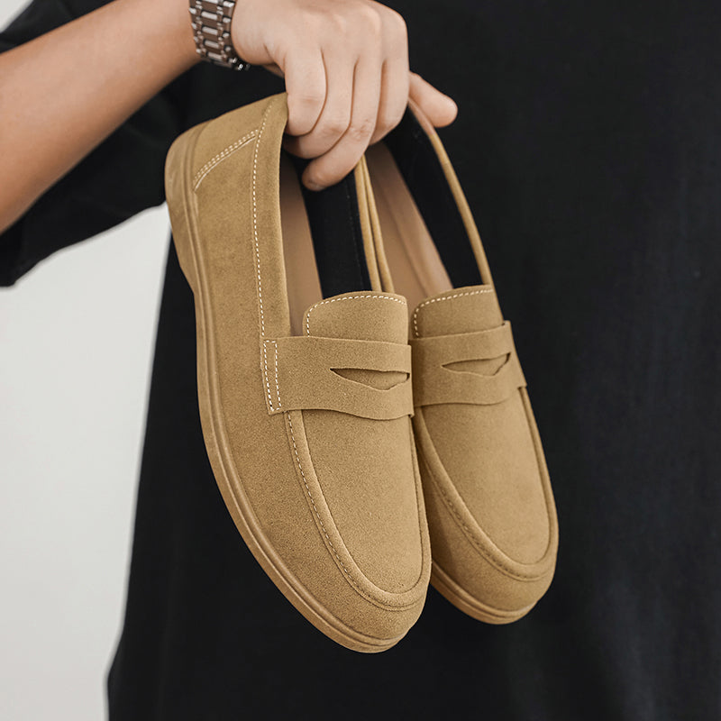 A hand holds a pair of tan suede loafers by the heels against a black t-shirt backdrop.