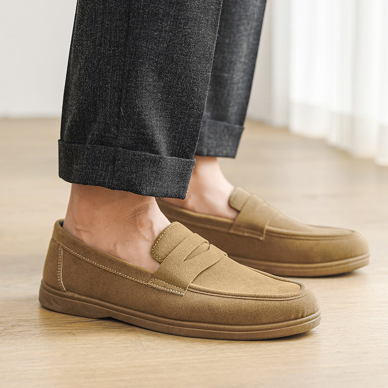 Close-up of a person's feet wearing tan suede loafers with cuffed gray dress trousers, standing on a wood floor.