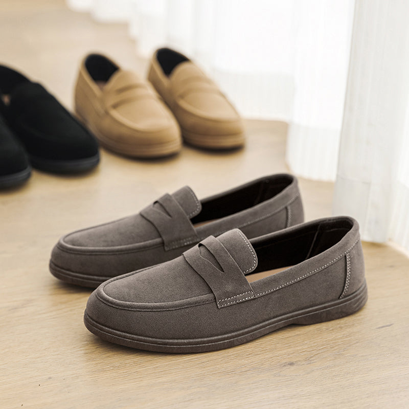 Gray suede penny loafers in the foreground with tan and black suede loafers lined up behind on a wooden floor beside a curtain.