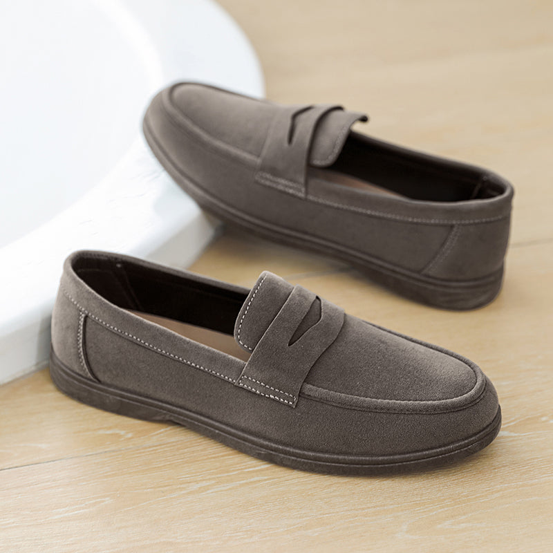 Pair of gray suede penny loafers resting on a wood floor against a curved white wall.