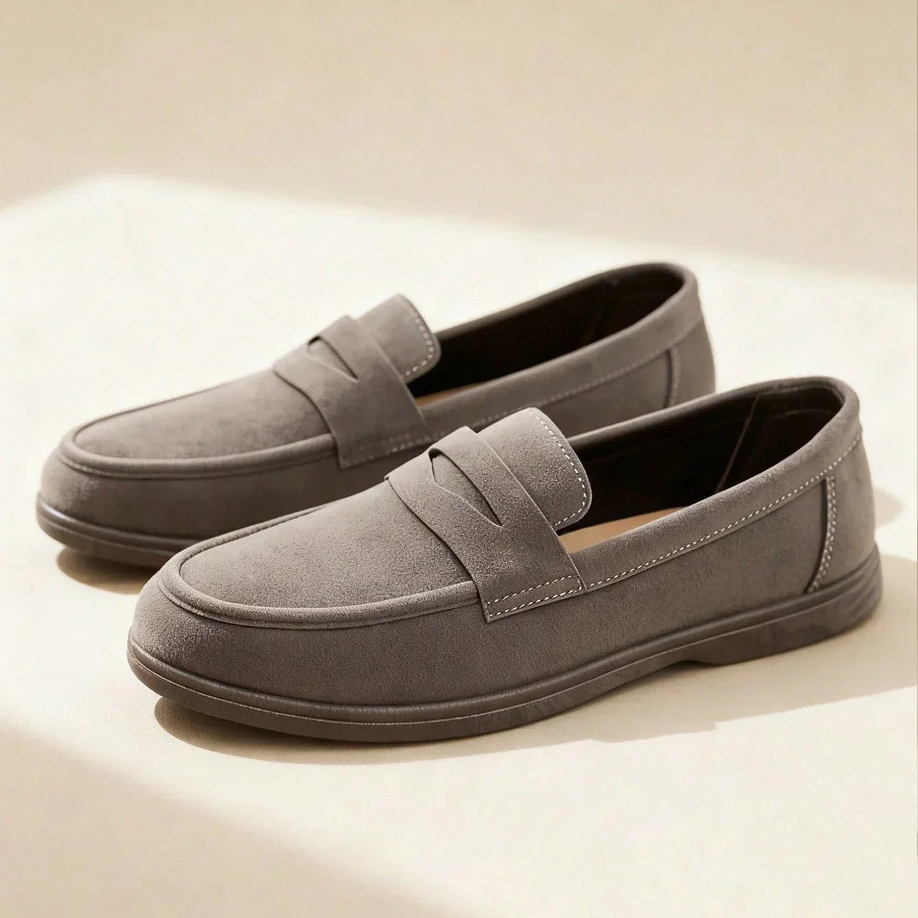 Pair of gray suede penny loafers on a beige surface under soft studio lighting.