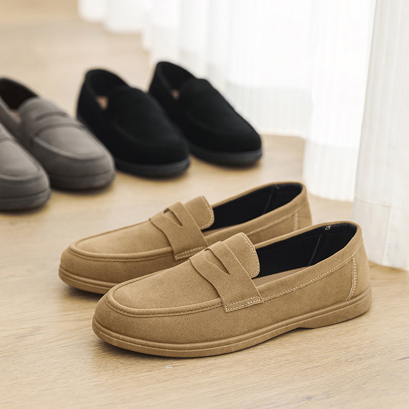 Tan suede penny loafers in the foreground with gray and black suede loafers lined up behind them on a wooden floor beside a curtain.