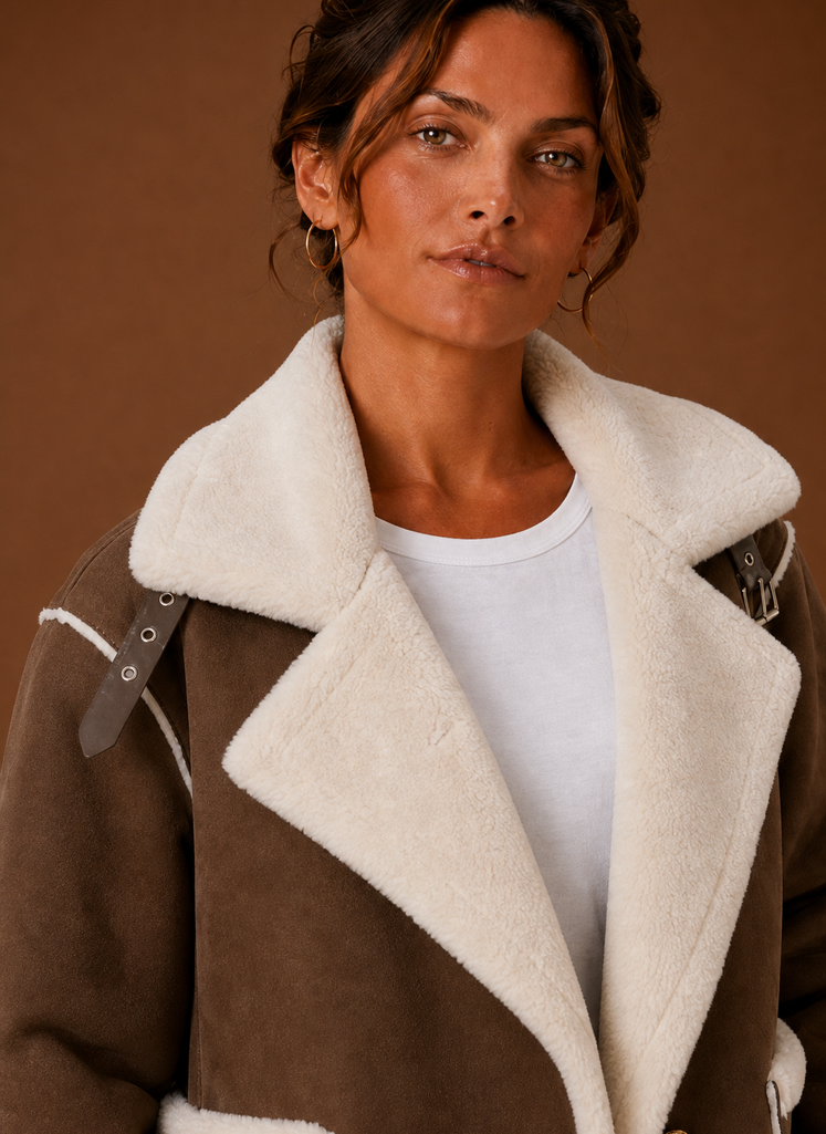 Brown suede-look aviator jacket with wide cream sherpa lapels and buckle strap details on a neutral background.