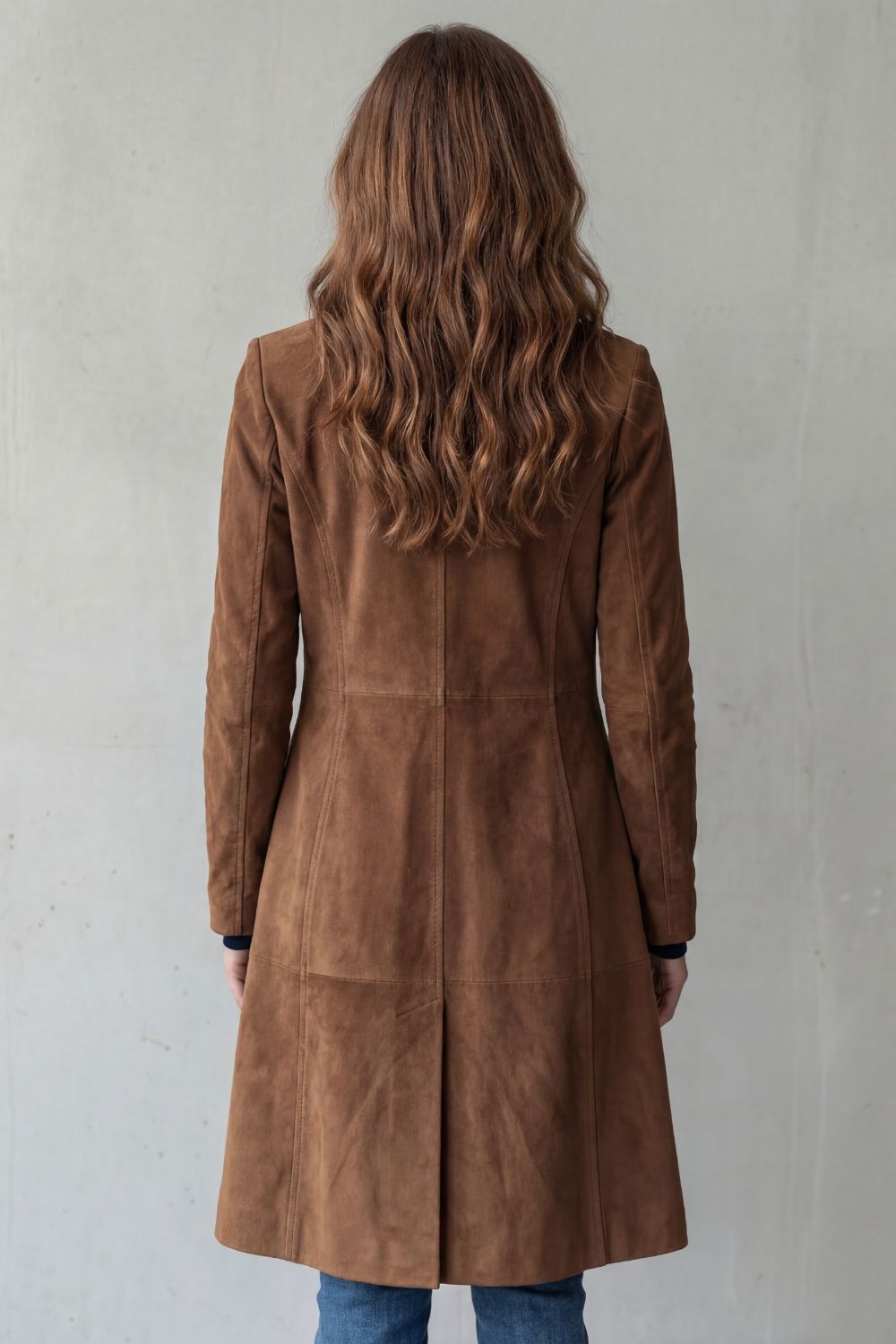 A woman wearing a brown suede longline coat with notch lapel and button-front closure, shown from the back