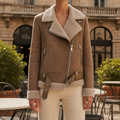 Brown suede moto jacket with cream teddy lining, silver zip hardware, and a belted waist in an outdoor cafe setting.