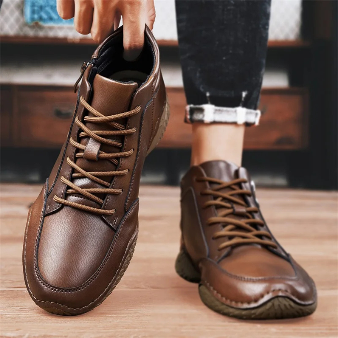 A person holding one brown leather zip-up ankle boot while wearing the other, cuffed jeans visible, standing on a wooden floor.