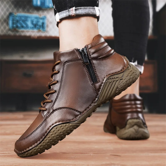 Close-up of a person holding one brown leather zip-up ankle boot while wearing the other, cuffed jeans visible, near a wooden dresser.