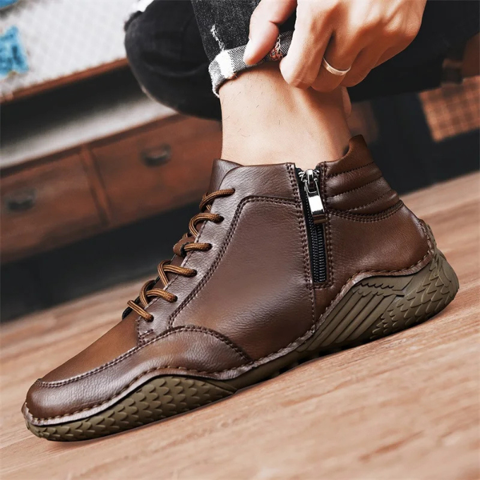 Close-up side profile of a person's ankle wearing a brown leather zip-up boot while adjusting a cuffed pant leg, on a wooden floor.