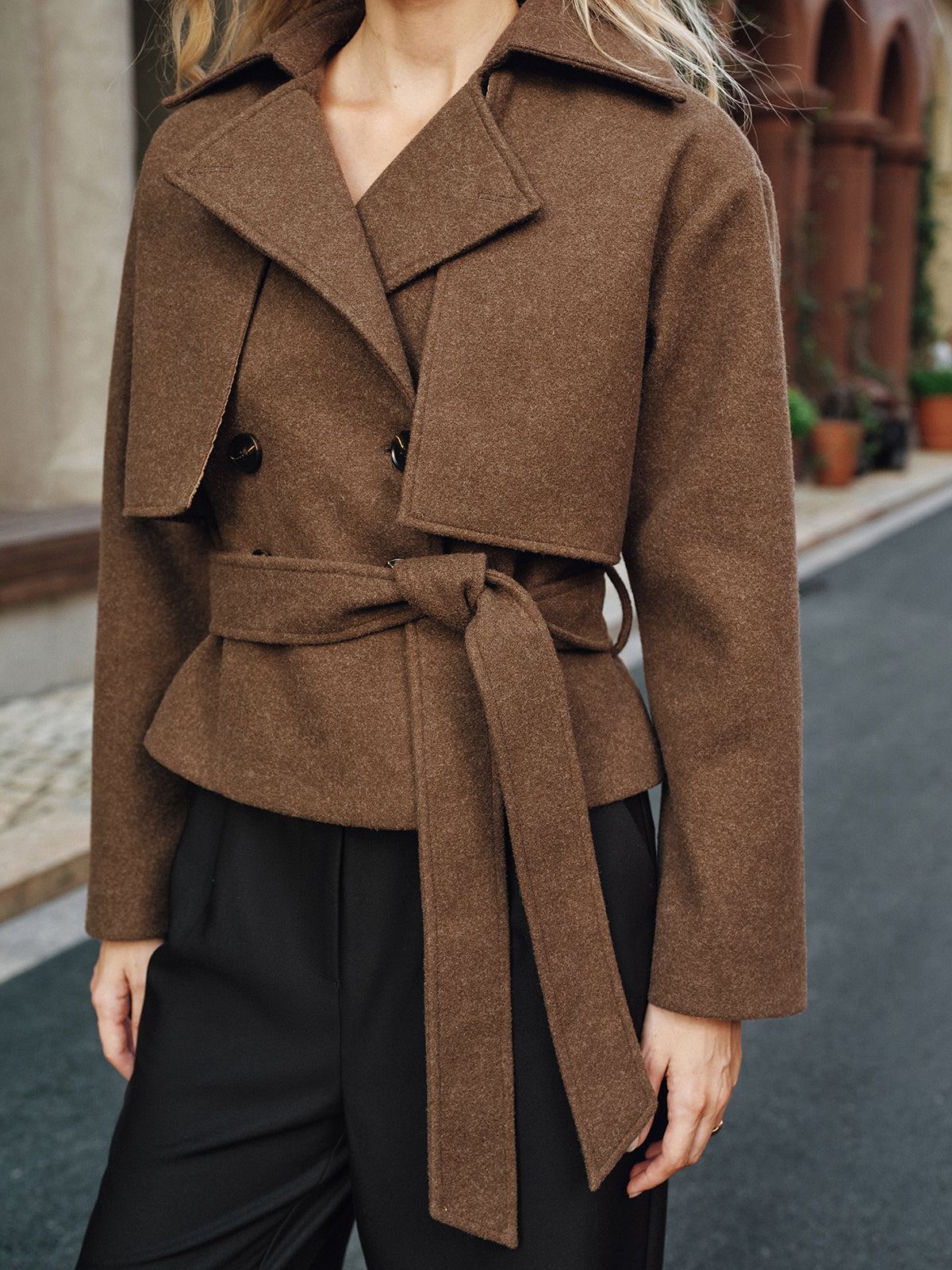 Close-up of a brown belted coat showing the wide lapel, double-breasted button closure, waist belt, and tailored construction.