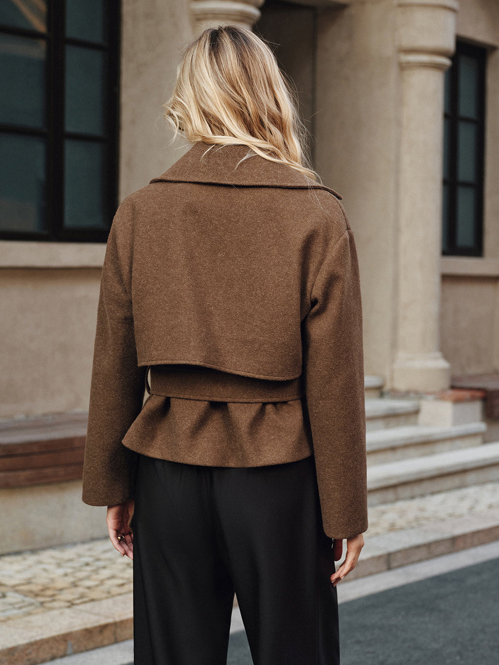 Back view of a woman wearing a brown belted coat with a structured silhouette and long sleeves.