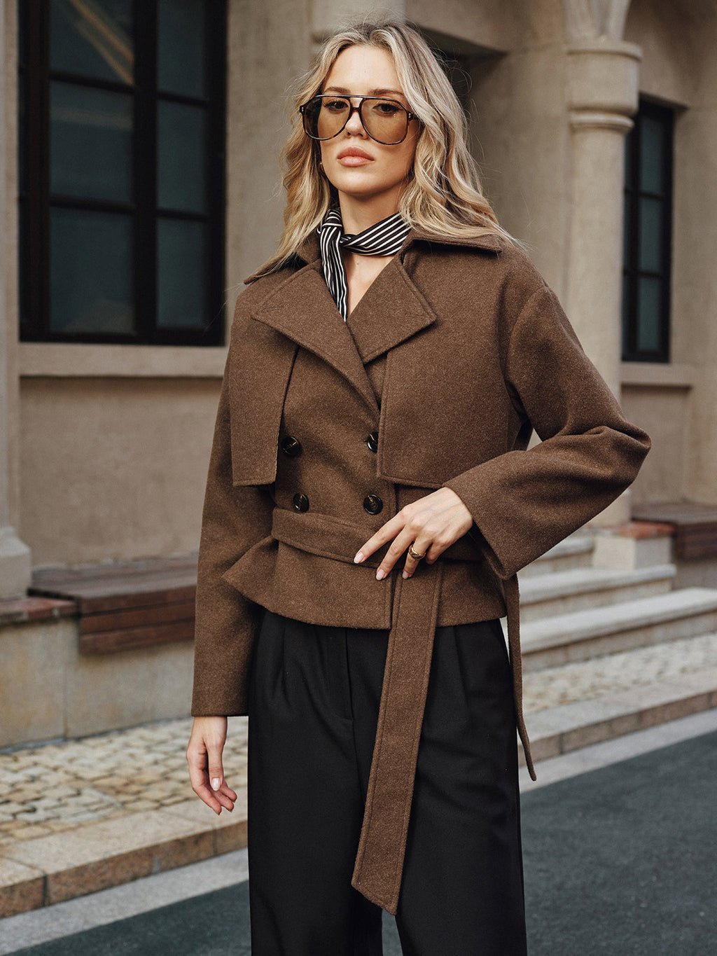 Woman wearing a brown belted coat with wide lapels, double-breasted buttons, and waist belt while standing outdoors.