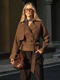 Woman wearing a brown belted coat with a wide notched lapel, double-breasted button closure, long sleeves, and tailored silhouette, paired with dark trousers and a brown handbag.