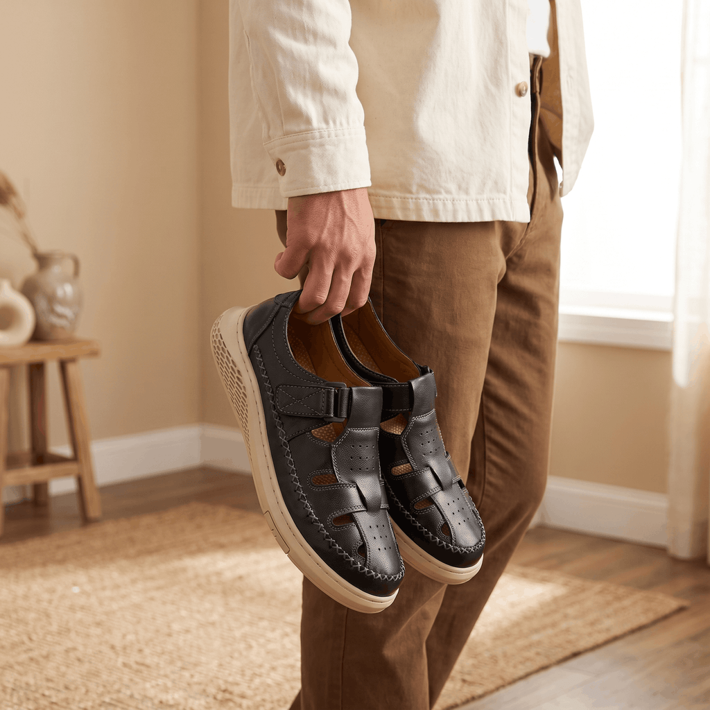 Man in cream jacket holding a pair of black leather Toby fisherman sandals, standing in a bright room on a jute rug near a window