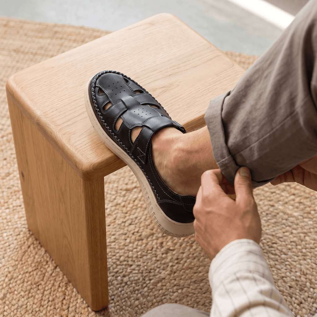 Man adjusting sock with foot resting on a wooden block wearing black leather Toby fisherman sandal, on a jute rug