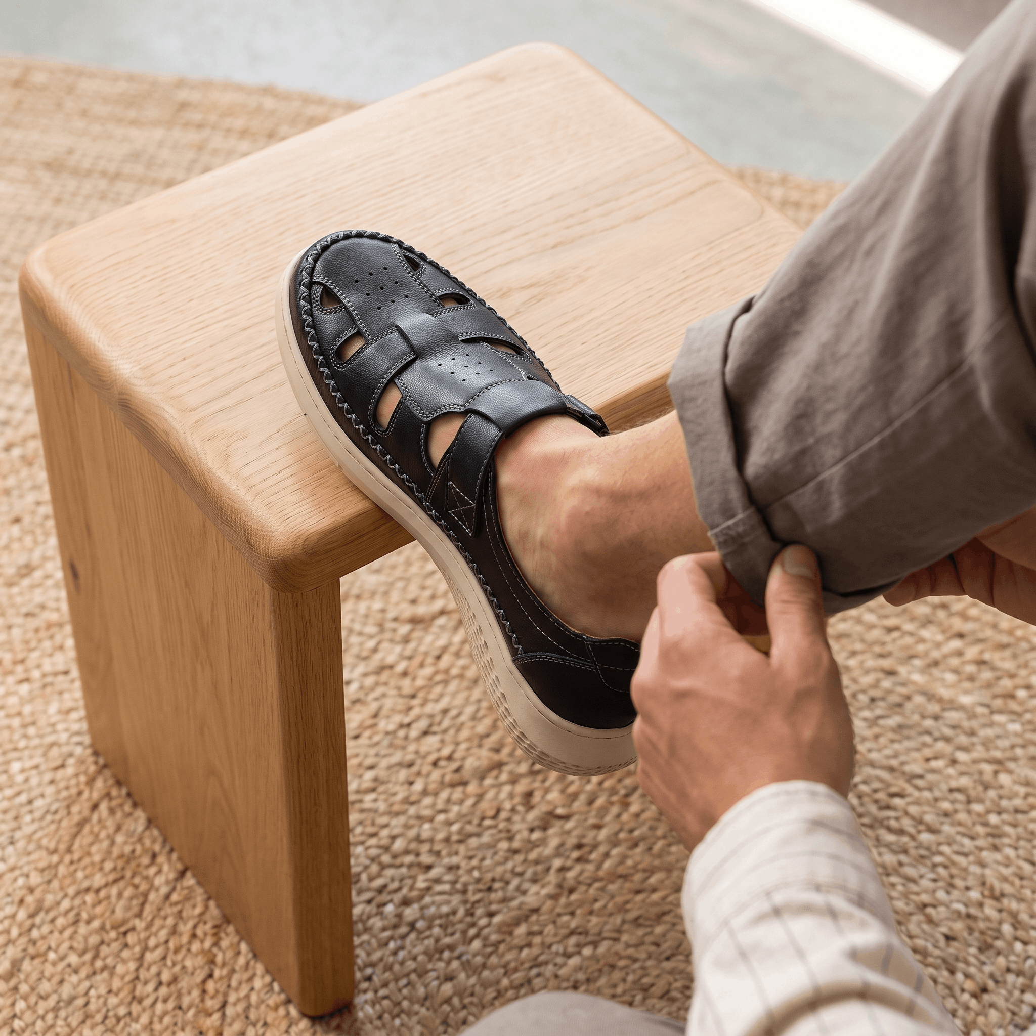 Man adjusting sock with foot resting on a wooden block wearing black leather Toby fisherman sandal, on a jute rug