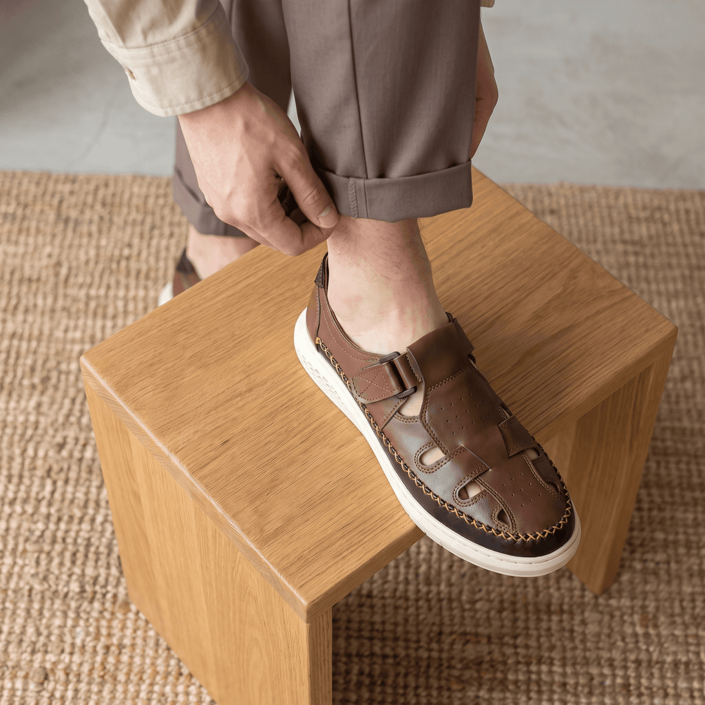 Man adjusting trouser cuff with foot resting on a wooden block, wearing brown leather Toby fisherman sandal over a jute rug