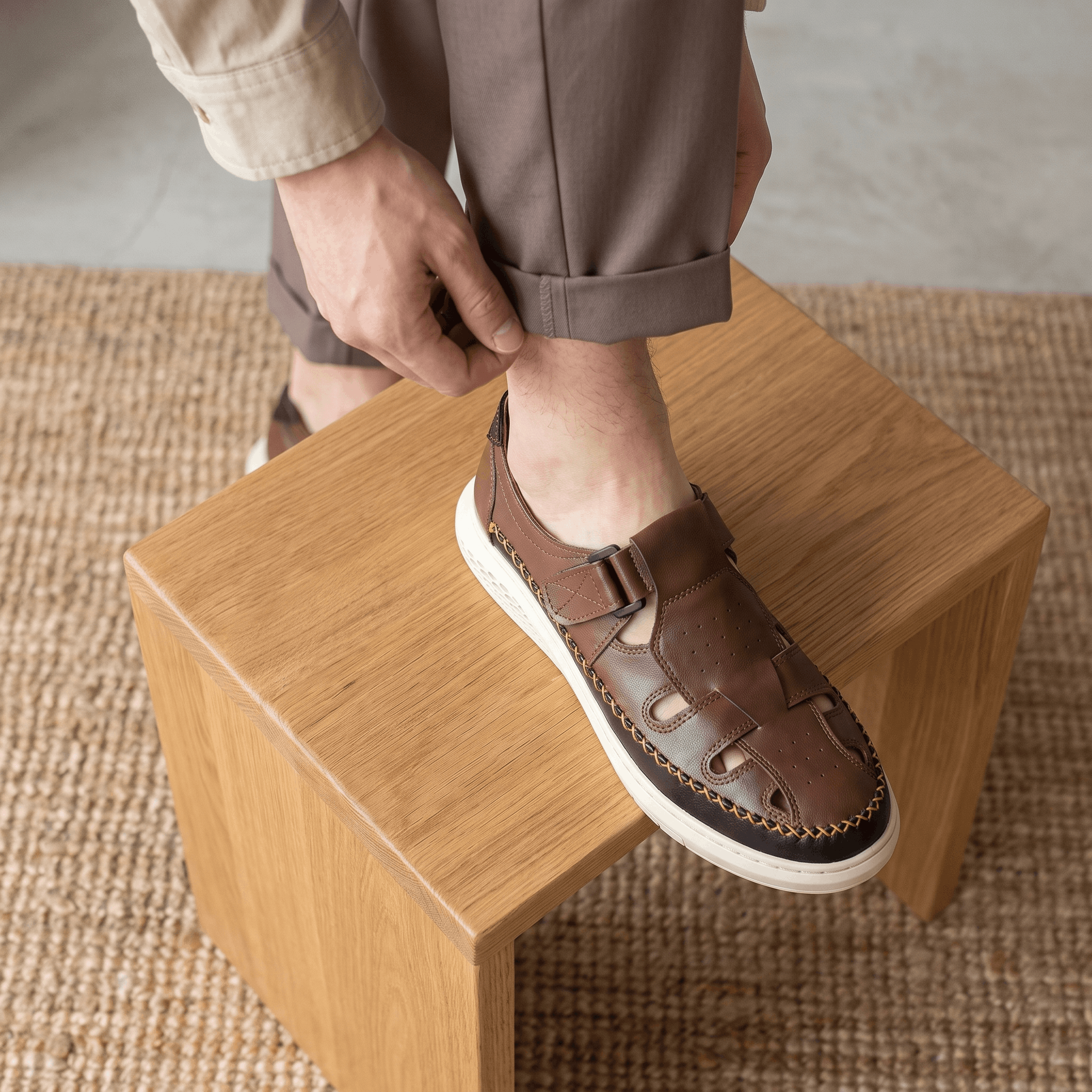 Man adjusting trouser cuff with foot resting on a wooden block, wearing brown leather Toby fisherman sandal over a jute rug