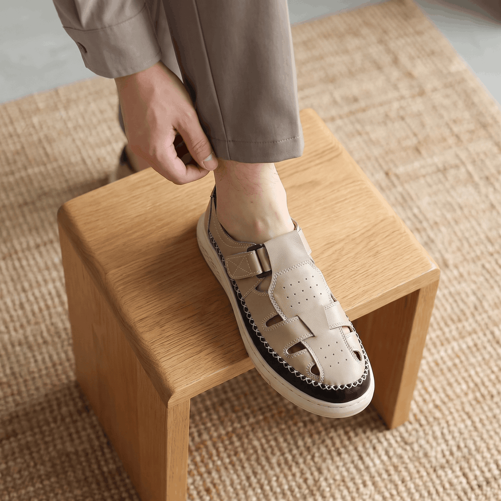 Man adjusting trouser cuff with foot resting on a wooden block, wearing beige leather Toby fisherman sandal over a jute rug