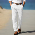 Man wearing white linen trousers with a relaxed straight-leg fit, pleated front, belt loops, and side pockets in a beachside setting.