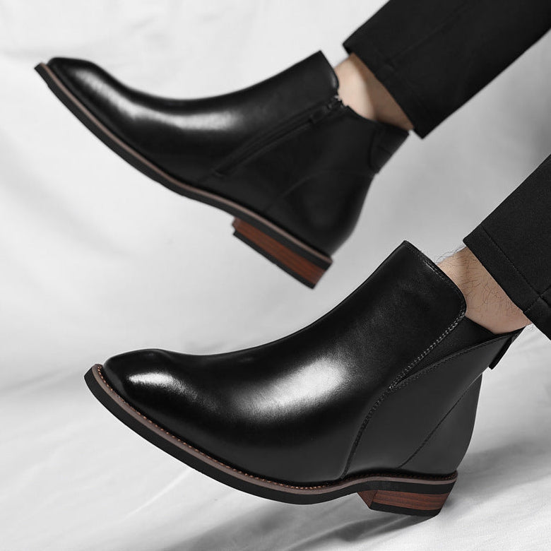 A model's ankles in black trousers wearing black leather Chelsea boots with side zippers, one foot lifted mid-step on a white sheet.