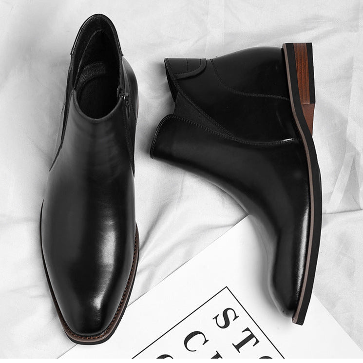 Overhead view of a pair of black leather Chelsea boots with side zippers, one tipped to show the sole, resting on a white sheet beside a magazine.