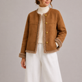 A woman wearing a brown vegan suede jacket with sherpa trim, gold-tone buttons, and a relaxed fit