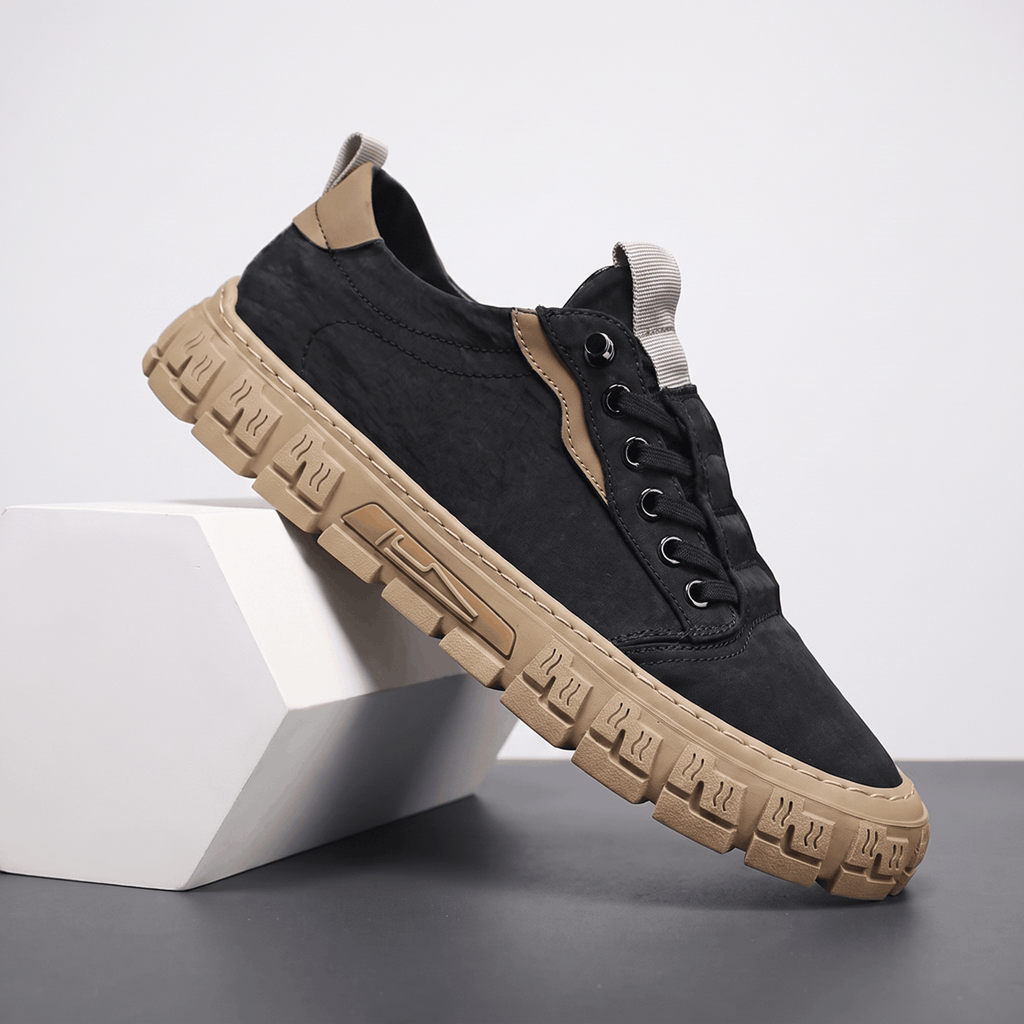 Black canvas sneaker with tan trim and tan treaded sole propped diagonally against a white geometric block in a studio setting