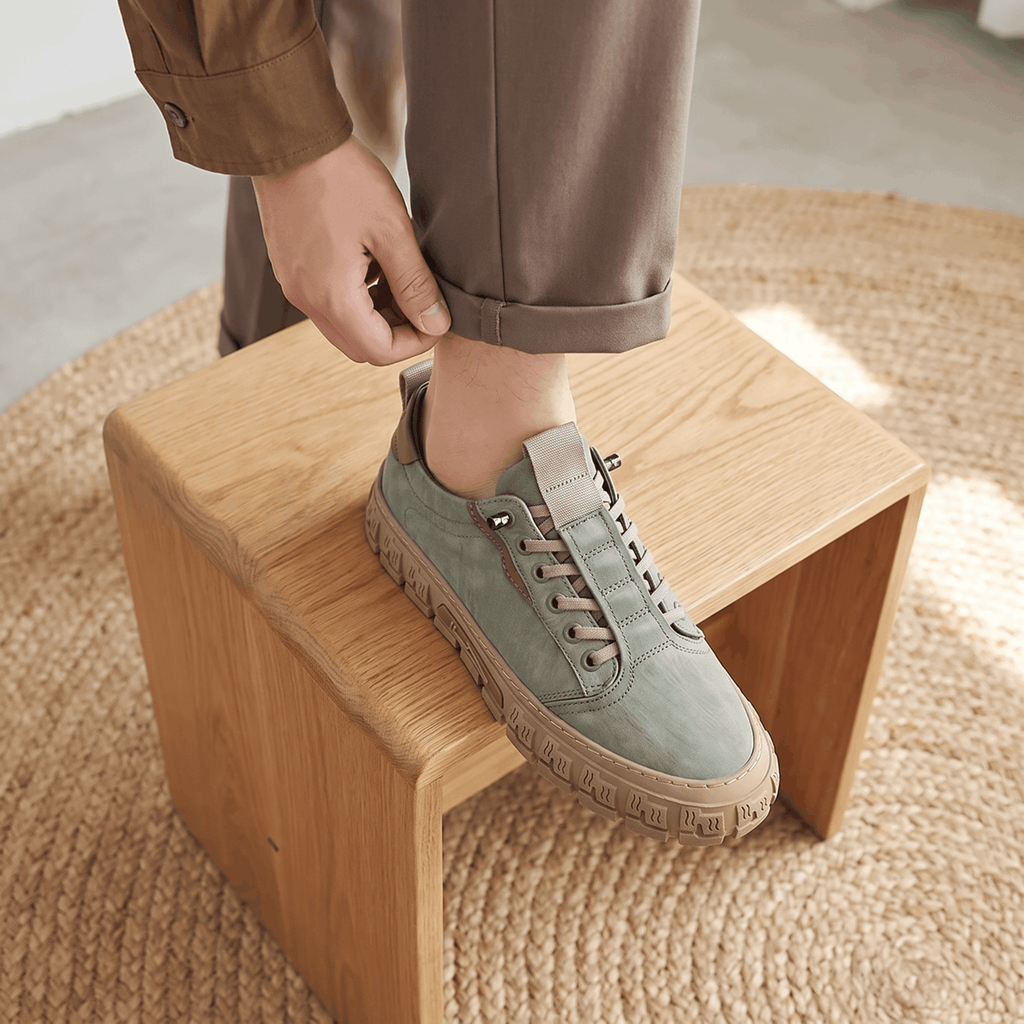 Person in brown trousers adjusting cuff with foot resting on a wooden block wearing a sage green suede sneaker, on a jute rug