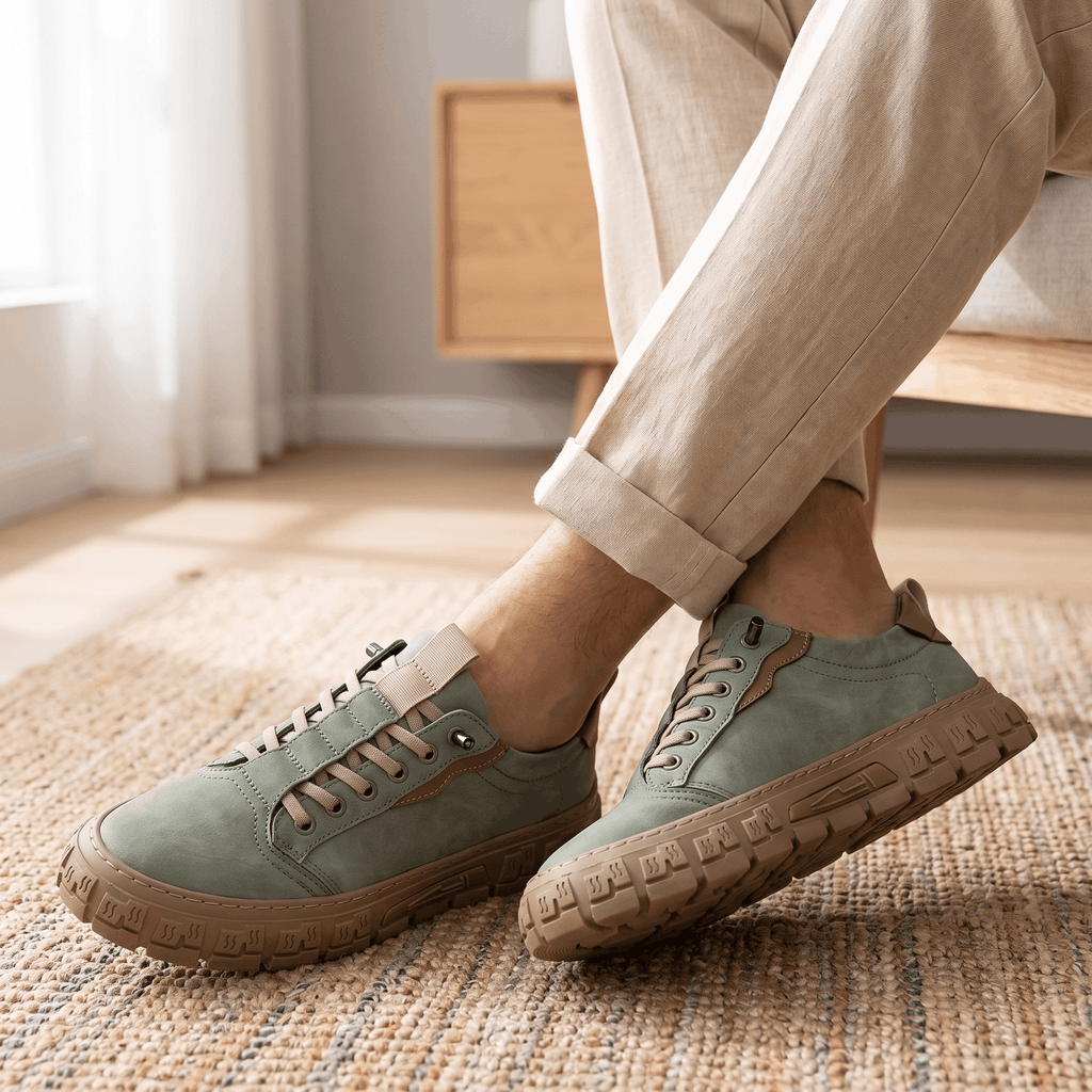 Close-up of legs in cuffed beige trousers wearing sage green suede sneakers, feet crossed on a woven rug in a sunlit room