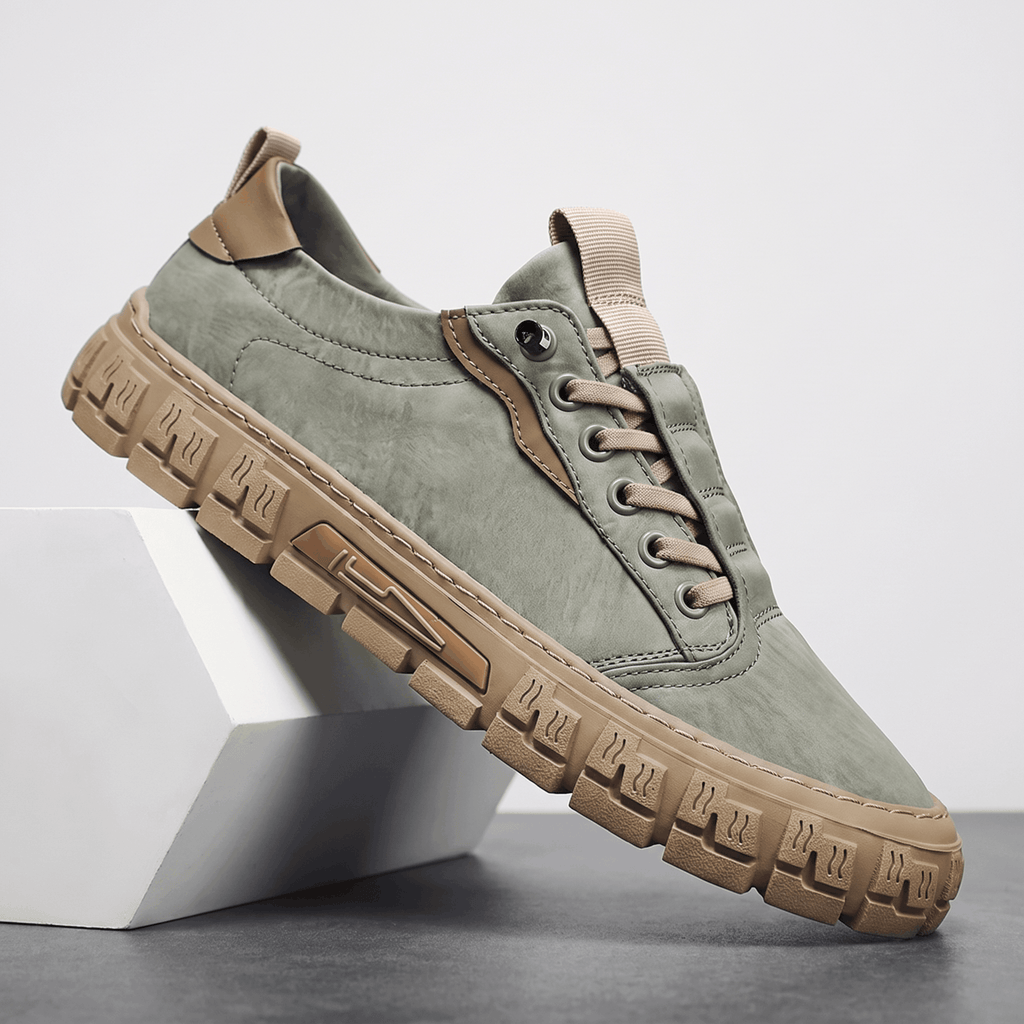 Sage green suede sneaker with tan leather trim and gum rubber sole propped diagonally against a white geometric block in a studio setting