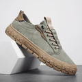 Sage green suede sneaker with tan leather trim and gum rubber sole propped diagonally against a white geometric block in a studio setting