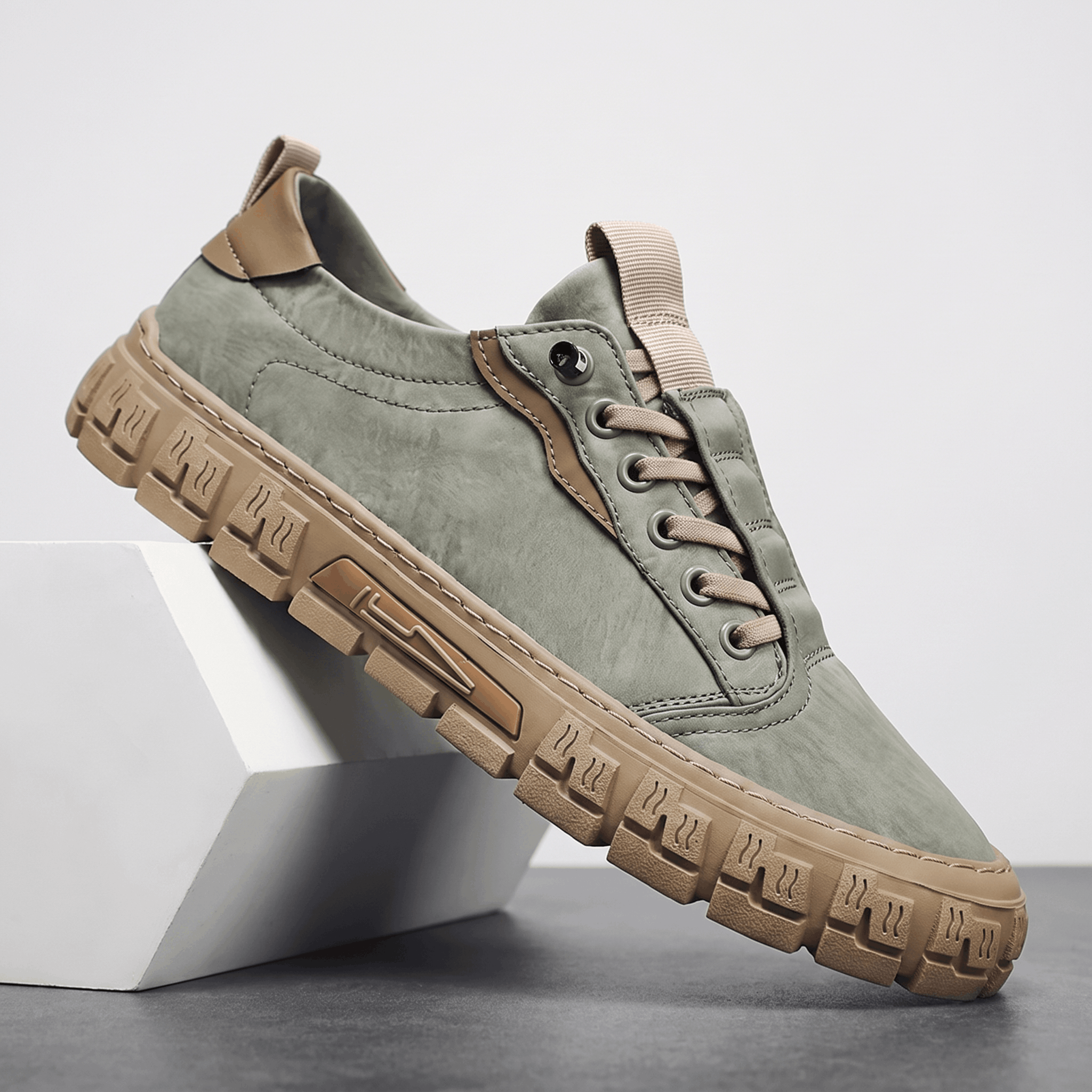 Sage green suede sneaker with tan leather trim and gum rubber sole propped diagonally against a white geometric block in a studio setting