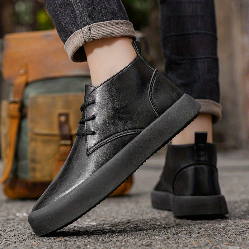 Close-up of a person's feet wearing black leather chukka-style boots and cuffed jeans, a backpack visible in the background on pavement.