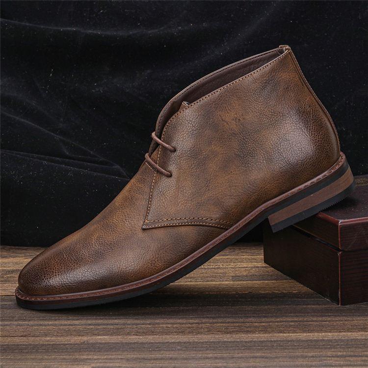 A single rustic brown leather chukka boot propped against a small wooden box, on a wooden floor with a dark draped backdrop.