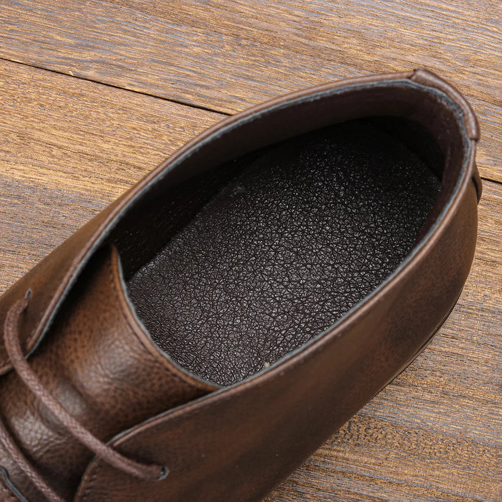 Close-up looking down into the opening of a dark brown leather chukka boot, showing the black interior lining, on a wooden surface.