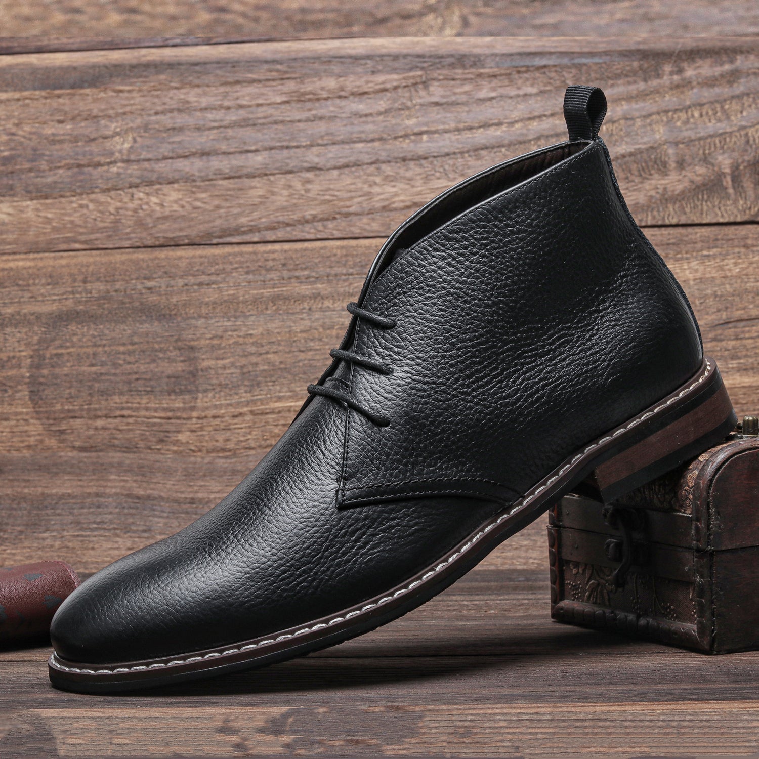 A single black pebbled leather chukka boot propped against a small wooden chest, on a wood-plank floor.