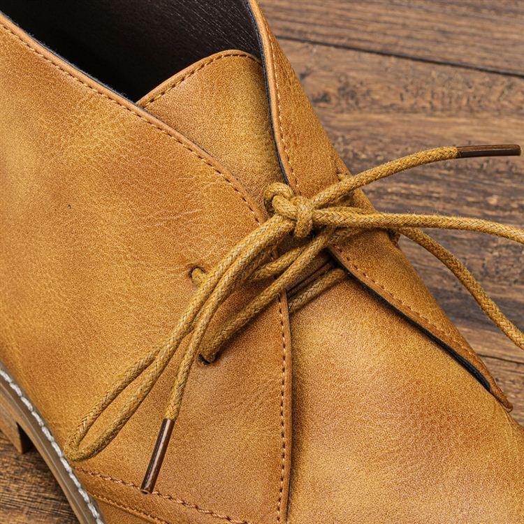 Extreme close-up of the tied laces and eyelets on a tan leather chukka boot, resting on a wooden surface.