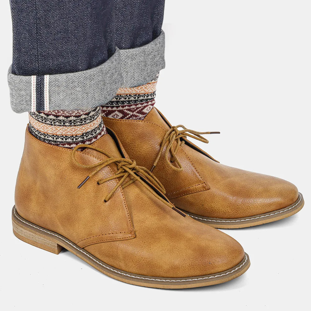 A model's feet in cuffed jeans and patterned socks wearing tan leather chukka boots, on a plain white background.