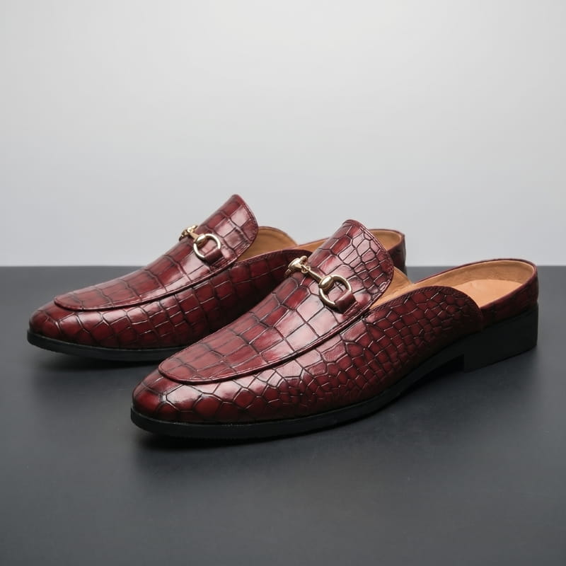 Pair of burgundy croc-embossed leather mule loafers with gold horsebit buckles arranged side by side on a gray backdrop